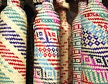 Colorful woven bottles feature intricate geometric patterns with a variety of vibrant colors including red, blue, purple, and natural straw. The craftsmanship suggests a blend of traditional weaving techniques with a modern aesthetic.