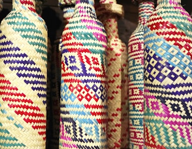 Colorful woven bottles feature intricate geometric patterns with a variety of vibrant colors including red, blue, purple, and natural straw. The craftsmanship suggests a blend of traditional weaving techniques with a modern aesthetic.