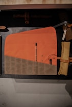 A neatly folded tool roll with orange and brown sections lies on a black surface. A pencil is placed within it, labeled with 'WINTER SESSION'. Adjacent to it, a tightly rolled tan fabric is secured with a leather strap.