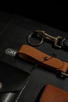 A close-up view of leather accessories including a brown leather strap with a metallic button and loop, alongside another metallic accessory on a dark surface.