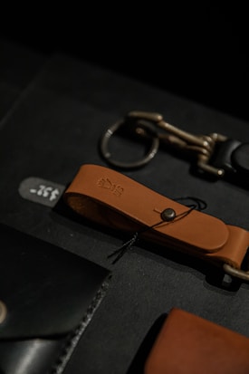 A close-up view of leather accessories including a brown leather strap with a metallic button and loop, alongside another metallic accessory on a dark surface.