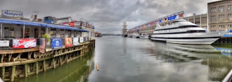 A waterfront scene with a restaurant on the left advertising various beer brands, adjacent to a large yacht named 'Spirit of Boston' docked on the right side. The setting appears to be a harbor or marina with a mix of cloudy skies and calm waters reflecting the buildings and boats.