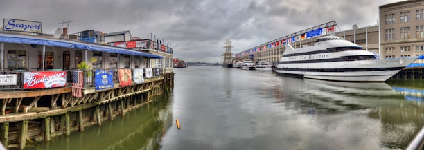 A waterfront scene with a restaurant on the left advertising various beer brands, adjacent to a large yacht named 'Spirit of Boston' docked on the right side. The setting appears to be a harbor or marina with a mix of cloudy skies and calm waters reflecting the buildings and boats.