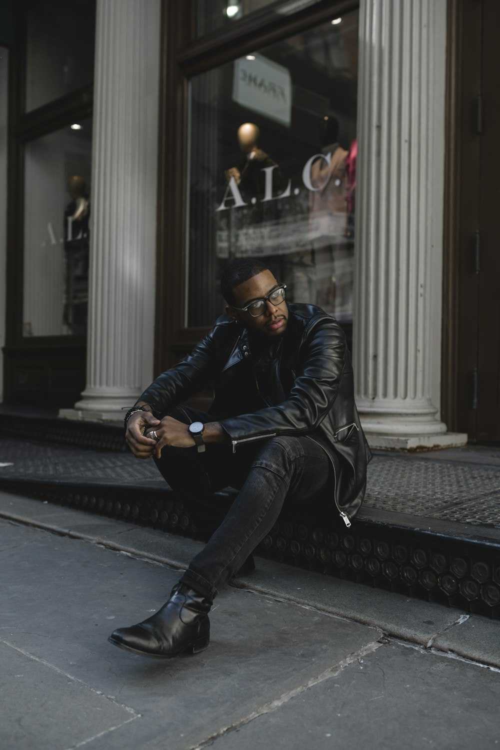 man in black leather jacket and black pants sitting on sidewalk during daytime