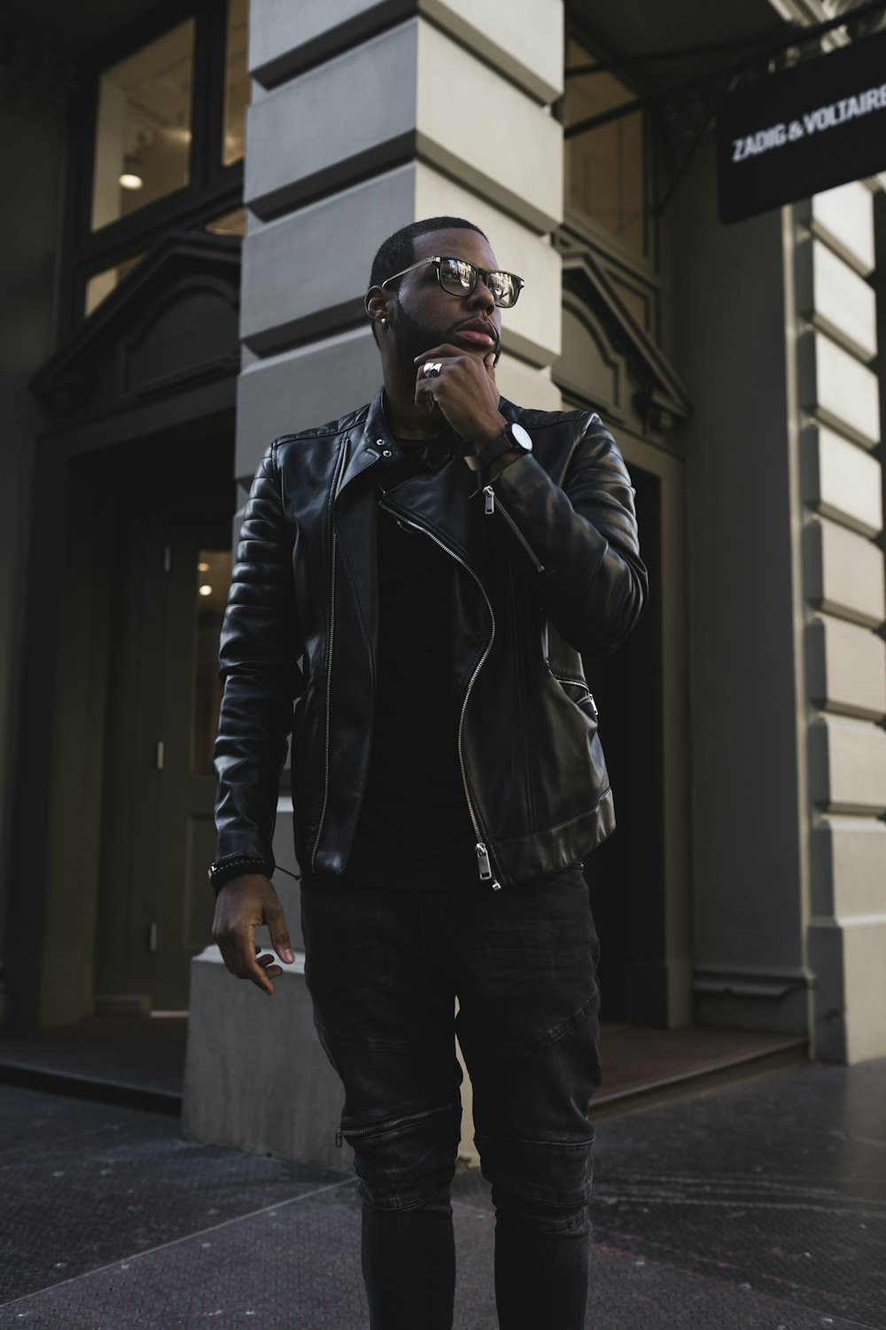 Hombre con chaqueta de cuero negro y pantalones negros con gafas de sol de pie al lado del edificio durante el día