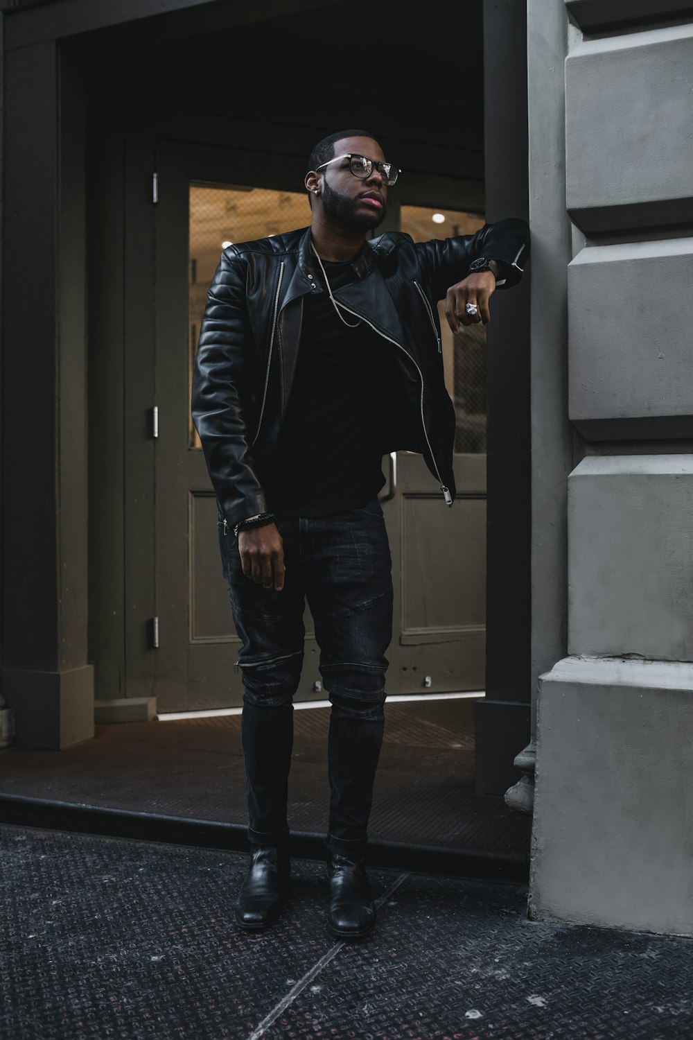 man in black zip up jacket and blue denim jeans standing beside gray concrete wall