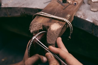 A craftsman skillfully holds a piece of jewelry, possibly a bracelet, adorned with small sparkling gems. The piece is positioned on a rustic wooden workbench, with various tools scattered around, indicating a workspace for jewelry making.