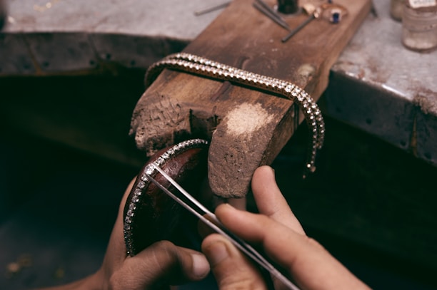 A craftsman skillfully holds a piece of jewelry, possibly a bracelet, adorned with small sparkling gems. The piece is positioned on a rustic wooden workbench, with various tools scattered around, indicating a workspace for jewelry making.