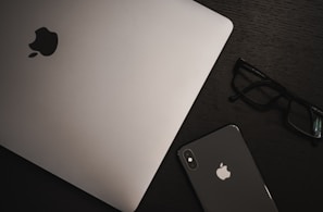 A MacBook and an iPhone with visible Apple logos are placed on a dark wooden surface, accompanied by a pair of black-framed glasses.