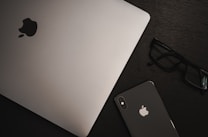 A MacBook and an iPhone with visible Apple logos are placed on a dark wooden surface, accompanied by a pair of black-framed glasses.