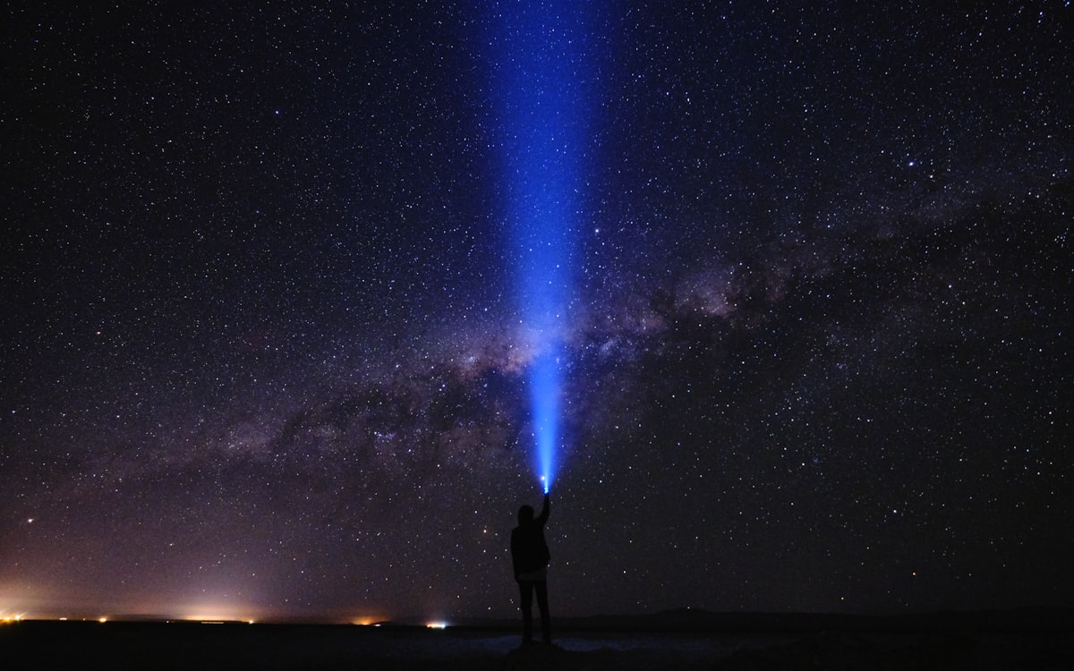 A person standing in the desert at night aiming a blue headlamp beam at the Milky Way, with town lights on the horizon