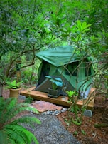 A green tent is set up on a wooden platform in a lush, green forested area. The scene is adorned with various potted plants, including ferns and broad-leafed plants. Wooden elements and natural stone decorations are present on the ground, creating a serene camping spot surrounded by dense greenery.