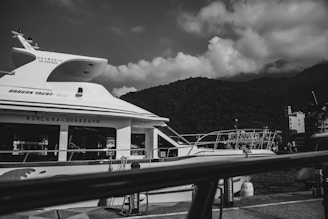 A large yacht docked at a marina, with a mountainous landscape in the background under a partly cloudy sky. The yacht has visible text on its side and appears to be well-maintained. The image is in black and white, giving it a classic feel.