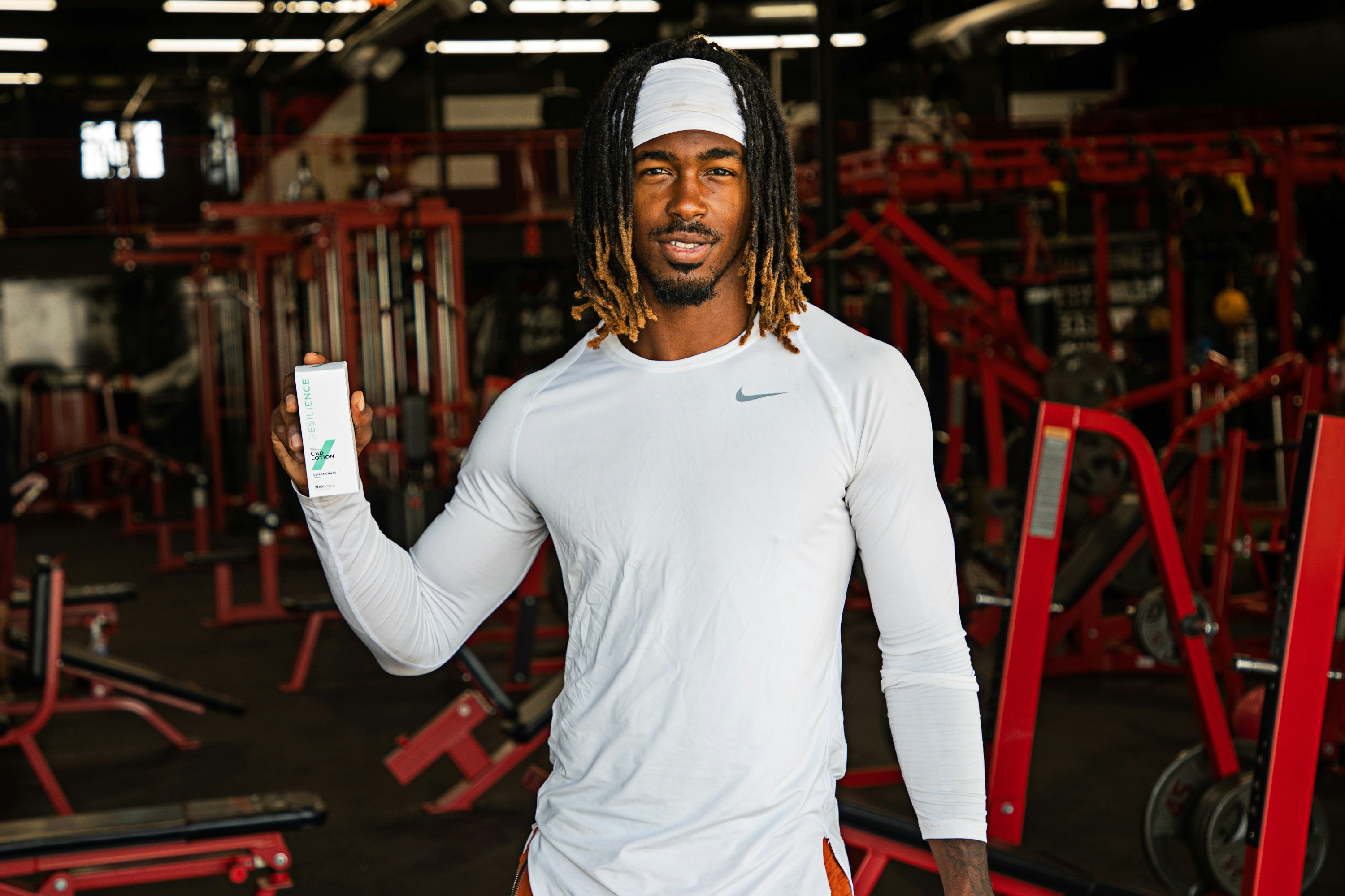 Male in white shirt holding CBD lotion with gym background.