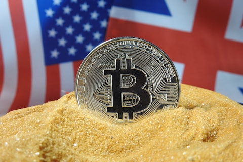 A physical Bitcoin coin partially buried in sand with the American and British flags blurred in the background.