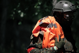 A person in a camouflage uniform and helmet is holding a high-visibility orange and black vest. The background is dark and blends into a forest setting. The figure is partially obscured by shadows, suggesting a tactical or covert environment.