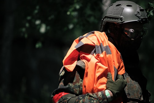 A person in a camouflage uniform and helmet is holding a high-visibility orange and black vest. The background is dark and blends into a forest setting. The figure is partially obscured by shadows, suggesting a tactical or covert environment.