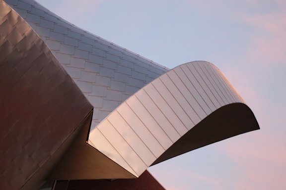 A section of a modern building is captured with a close-up view of its metallic architectural design. The structure features overlapping metal panels and a curved roof element, displaying smooth, reflective surfaces.