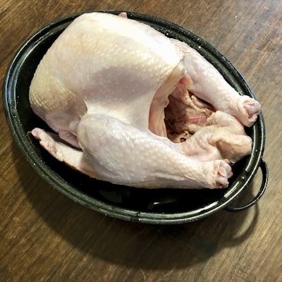 A raw whole chicken is placed in a black roasting pan on a wooden surface. The chicken's skin is pale, and it is unseasoned, ready for preparation.