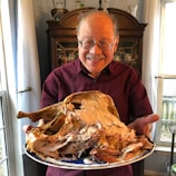 An older man with glasses and a mustache is smiling and holding a large platter of carved turkey. He is wearing a maroon patterned shirt and standing indoors with a wooden cabinet and large window in the background.
