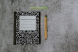 A black and white composition notebook is placed on a speckled gray surface alongside a green pen and two yellow pencils with pink erasers.