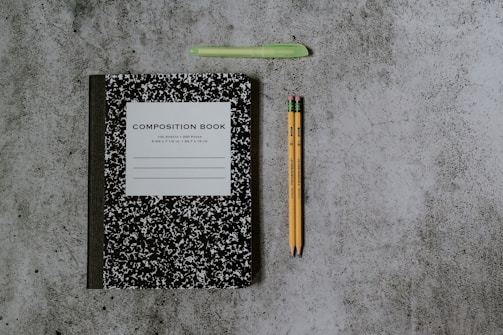 A black and white composition notebook is placed on a speckled gray surface alongside a green pen and two yellow pencils with pink erasers.