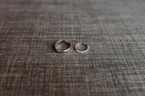 Two rings are placed side by side on a textured grey fabric surface. One ring is a plain band, while the other appears to have a row of small stones or diamonds.