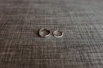 Two rings are placed side by side on a textured grey fabric surface. One ring is a plain band, while the other appears to have a row of small stones or diamonds.