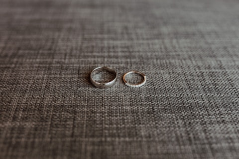 Two rings are placed side by side on a textured grey fabric surface. One ring is a plain band, while the other appears to have a row of small stones or diamonds.