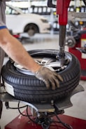 A person is using a tire changing machine to remove or fit a tire onto a wheel. The gloved hand is holding the tire in place while the machine's arm is assisting with the process. In the background, there is a white car and shelves filled with what appear to be containers or boxes.