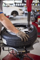 A person is using a tire changing machine to remove or fit a tire onto a wheel. The gloved hand is holding the tire in place while the machine's arm is assisting with the process. In the background, there is a white car and shelves filled with what appear to be containers or boxes.