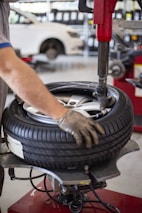 A person is using a tire changing machine to remove or fit a tire onto a wheel. The gloved hand is holding the tire in place while the machine's arm is assisting with the process. In the background, there is a white car and shelves filled with what appear to be containers or boxes.