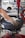 A person is using a tire changing machine to remove or fit a tire onto a wheel. The gloved hand is holding the tire in place while the machine's arm is assisting with the process. In the background, there is a white car and shelves filled with what appear to be containers or boxes.