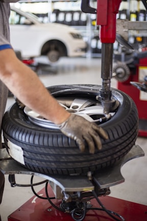 A person is using a tire changing machine to remove or fit a tire onto a wheel. The gloved hand is holding the tire in place while the machine's arm is assisting with the process. In the background, there is a white car and shelves filled with what appear to be containers or boxes.