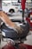 A person is using a tire changing machine to remove or fit a tire onto a wheel. The gloved hand is holding the tire in place while the machine's arm is assisting with the process. In the background, there is a white car and shelves filled with what appear to be containers or boxes.
