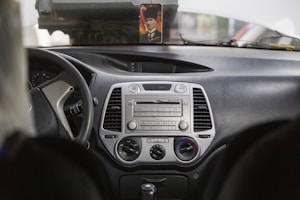The interior of a car is shown with a focus on the dashboard and console area. The dashboard features a central stereo system with various buttons and a display screen. Air vents are located on either side of the stereo system. The steering wheel is on the left side, and part of it is visible. A small, framed photo is placed on the dashboard. The overall car interior appears to be in neutral colors, primarily black and grey.