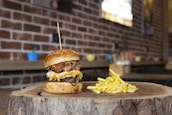 A gourmet cheeseburger with a thick beef patty, melted cheese, crispy bacon, and fried onions is served on a wooden slab, accompanied by a portion of golden fries. The setting features a rustic brick wall background and colorful potted plants in soft focus.