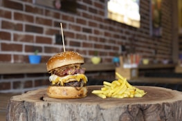 A gourmet cheeseburger with a thick beef patty, melted cheese, crispy bacon, and fried onions is served on a wooden slab, accompanied by a portion of golden fries. The setting features a rustic brick wall background and colorful potted plants in soft focus.