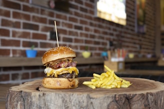A gourmet cheeseburger with a thick beef patty, melted cheese, crispy bacon, and fried onions is served on a wooden slab, accompanied by a portion of golden fries. The setting features a rustic brick wall background and colorful potted plants in soft focus.