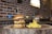 A gourmet cheeseburger with a thick beef patty, melted cheese, crispy bacon, and fried onions is served on a wooden slab, accompanied by a portion of golden fries. The setting features a rustic brick wall background and colorful potted plants in soft focus.