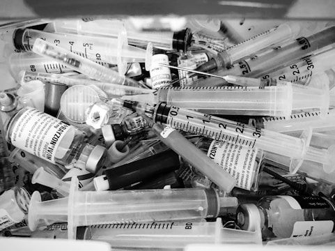 A collection of medical syringes, vials, and needles are piled together in a chaotic manner. The syringes vary in sizes, and some of the vials have labels with text on them. The overall scene appears cluttered and disorganized.