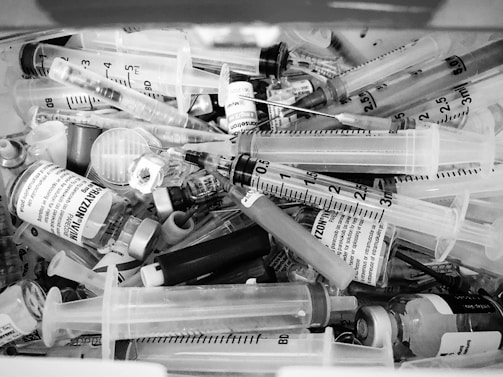 A collection of medical syringes, vials, and needles are piled together in a chaotic manner. The syringes vary in sizes, and some of the vials have labels with text on them. The overall scene appears cluttered and disorganized.