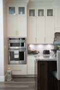 A modern kitchen with cream-colored cabinets featuring upper glass doors holding various containers and a decorative jug. The kitchen includes a built-in double oven with a towel hanging on the handle, adjacent to a countertop displaying a black toaster and a decorative bowl of greenery. The backsplash consists of light gray tiles, and there's a lantern on the floor. A part of an island with a marble countertop and dark wooden base is visible in the foreground.