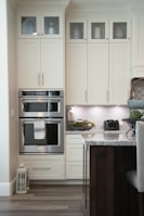 A modern kitchen with cream-colored cabinets featuring upper glass doors holding various containers and a decorative jug. The kitchen includes a built-in double oven with a towel hanging on the handle, adjacent to a countertop displaying a black toaster and a decorative bowl of greenery. The backsplash consists of light gray tiles, and there's a lantern on the floor. A part of an island with a marble countertop and dark wooden base is visible in the foreground.