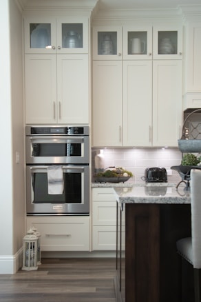 A modern kitchen with cream-colored cabinets featuring upper glass doors holding various containers and a decorative jug. The kitchen includes a built-in double oven with a towel hanging on the handle, adjacent to a countertop displaying a black toaster and a decorative bowl of greenery. The backsplash consists of light gray tiles, and there's a lantern on the floor. A part of an island with a marble countertop and dark wooden base is visible in the foreground.
