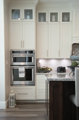 A modern kitchen with cream-colored cabinets featuring upper glass doors holding various containers and a decorative jug. The kitchen includes a built-in double oven with a towel hanging on the handle, adjacent to a countertop displaying a black toaster and a decorative bowl of greenery. The backsplash consists of light gray tiles, and there's a lantern on the floor. A part of an island with a marble countertop and dark wooden base is visible in the foreground.