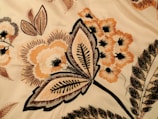 Intricate floral embroidery with detailed patterns in shades of orange, beige, and black. Large leaves and blooming flowers create a vintage and artistic appearance.