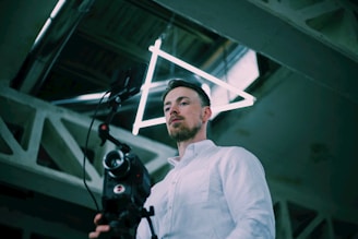 A man in a white shirt is holding a camera with professional equipment, standing in an industrial-looking setting with geometric metal beams. A bright neon triangular light fixture is positioned above his head.