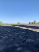 Close-up of an asphalt road with a clear blue sky in the background. The focus is on the textured surface of the road, while the background is blurred, showing vague shapes of trees or bushes along the horizon.
