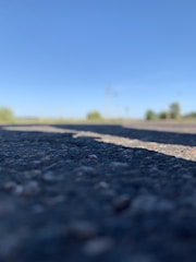 Close-up of an asphalt road with a clear blue sky in the background. The focus is on the textured surface of the road, while the background is blurred, showing vague shapes of trees or bushes along the horizon.